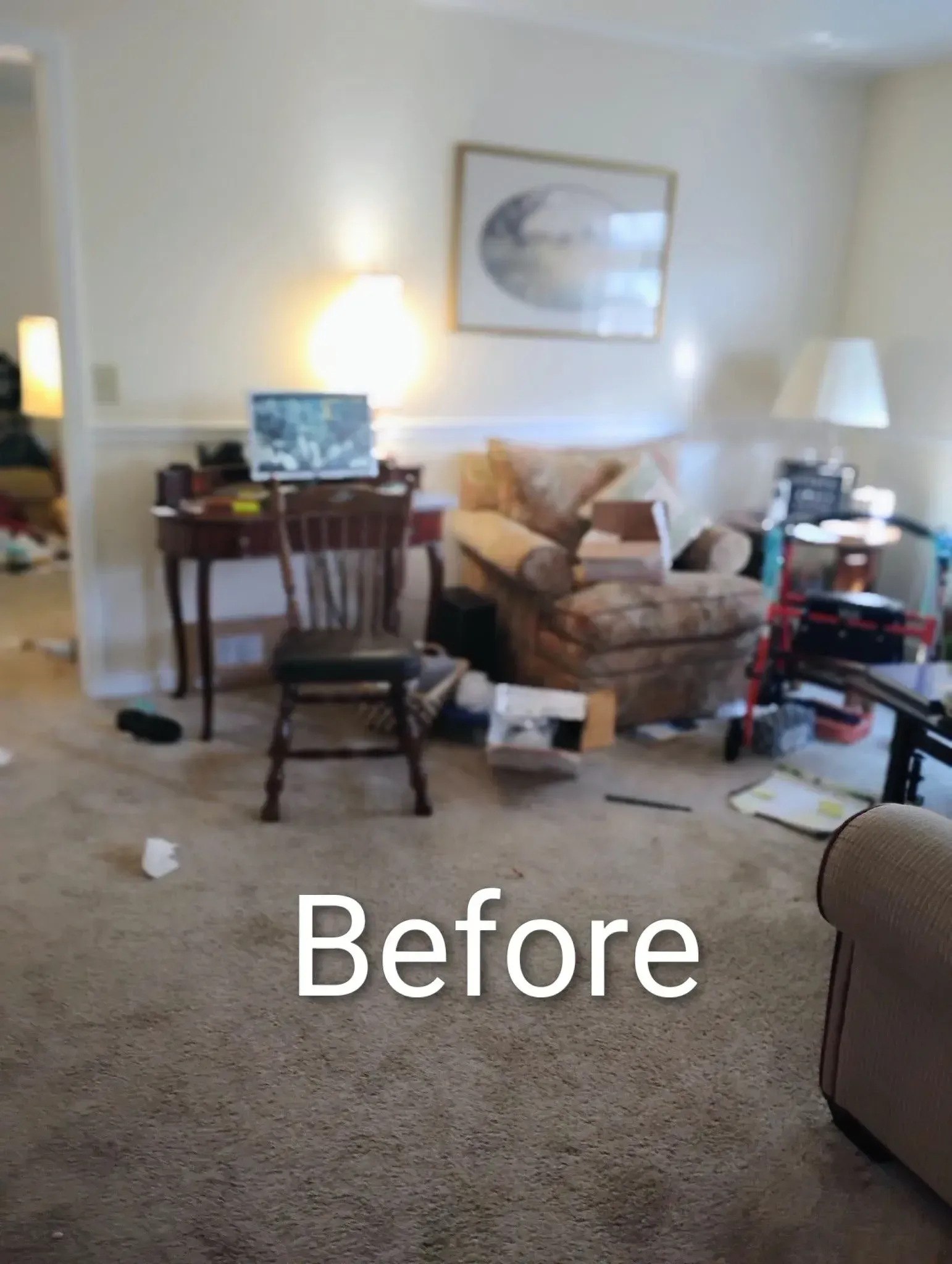 Cluttered living room with beige carpet, a desk, armchair, lamps, and various items scattered around.