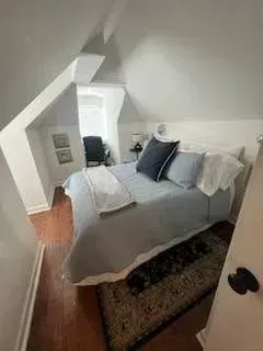 Bedroom with angled ceilings, bed, rug, dark chair in window light.