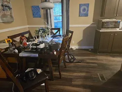 Dining room with messy table, wooden chairs, and kitchen cabinets. Brown flooring, cream walls, and natural light.