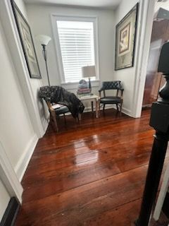 A hallway with wooden floors , chairs , a lamp and a window.