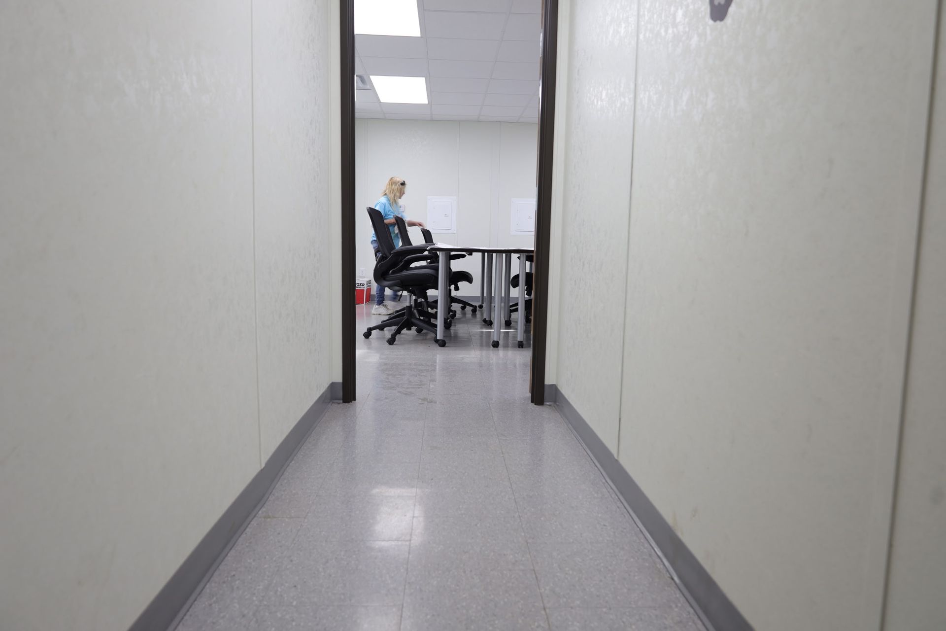 Hallway leading to a room with a person seated at a table. White walls and gray floor.