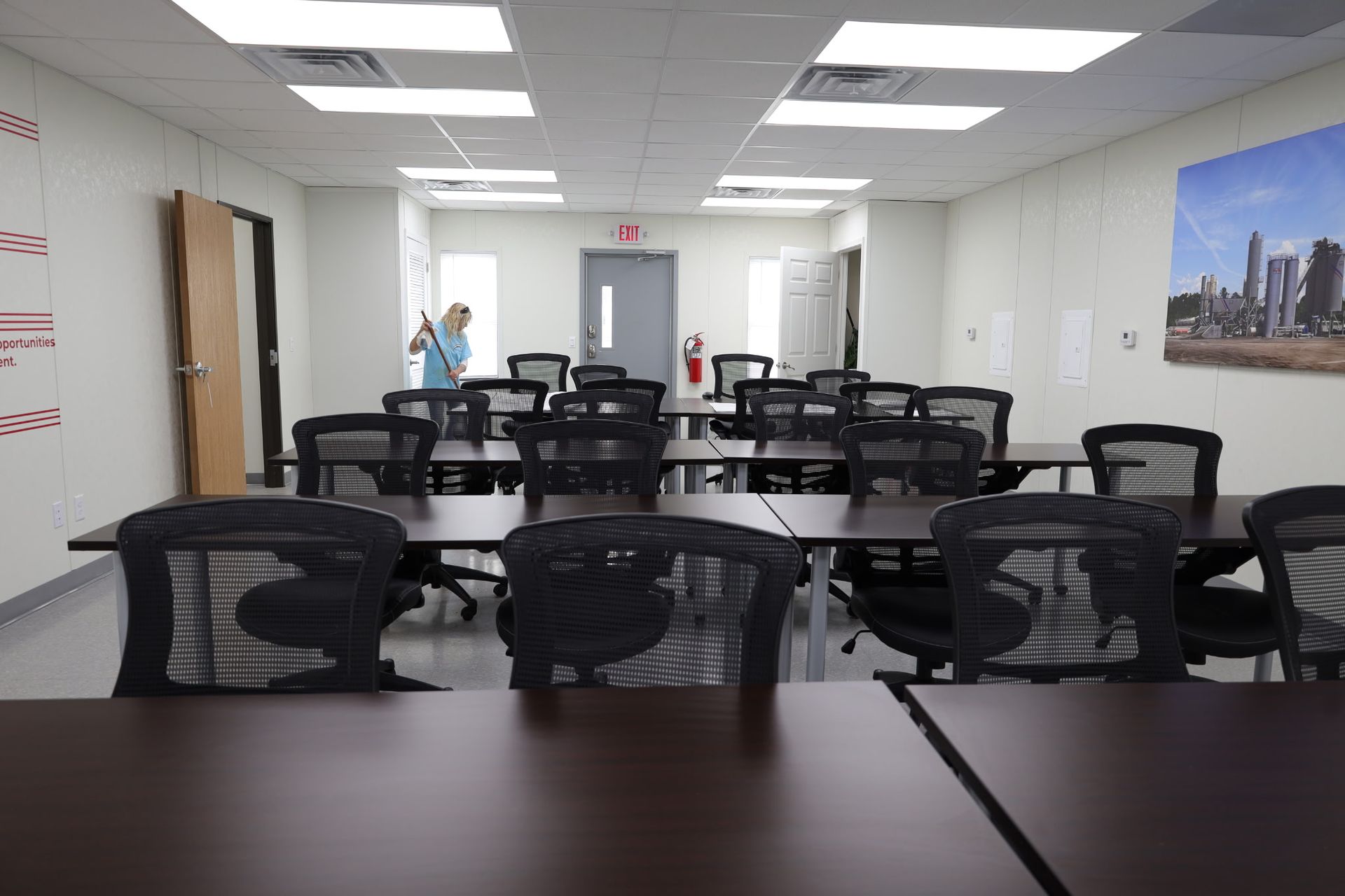 Empty classroom with rows of desks, black chairs, and a person by the doorway.