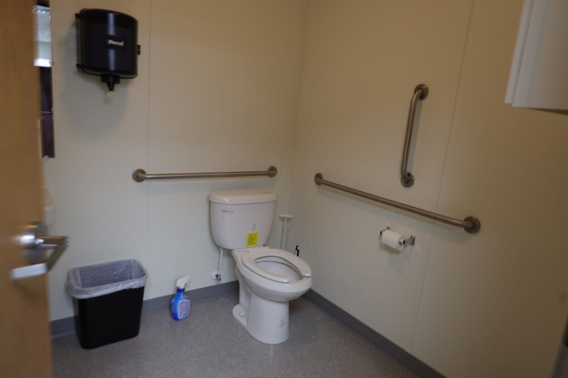 Accessible restroom with grab bars, toilet, paper towel dispenser, trash can, and cleaning spray.