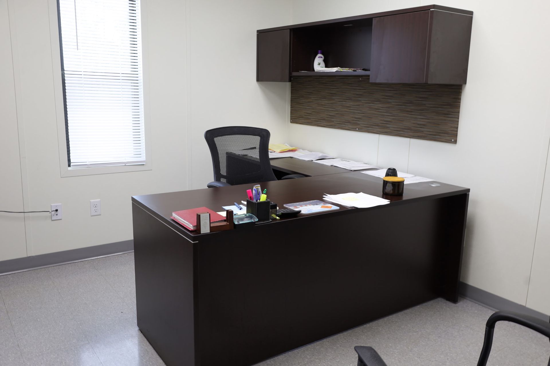 An office with a dark brown L-shaped desk, black chair, and overhead cabinet against a beige wall.