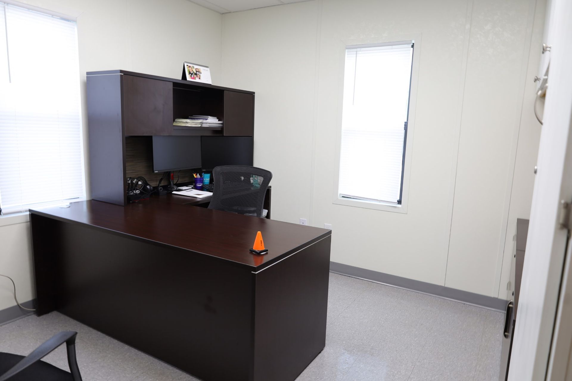 Office with a dark brown desk, black chair, two windows, and beige walls.
