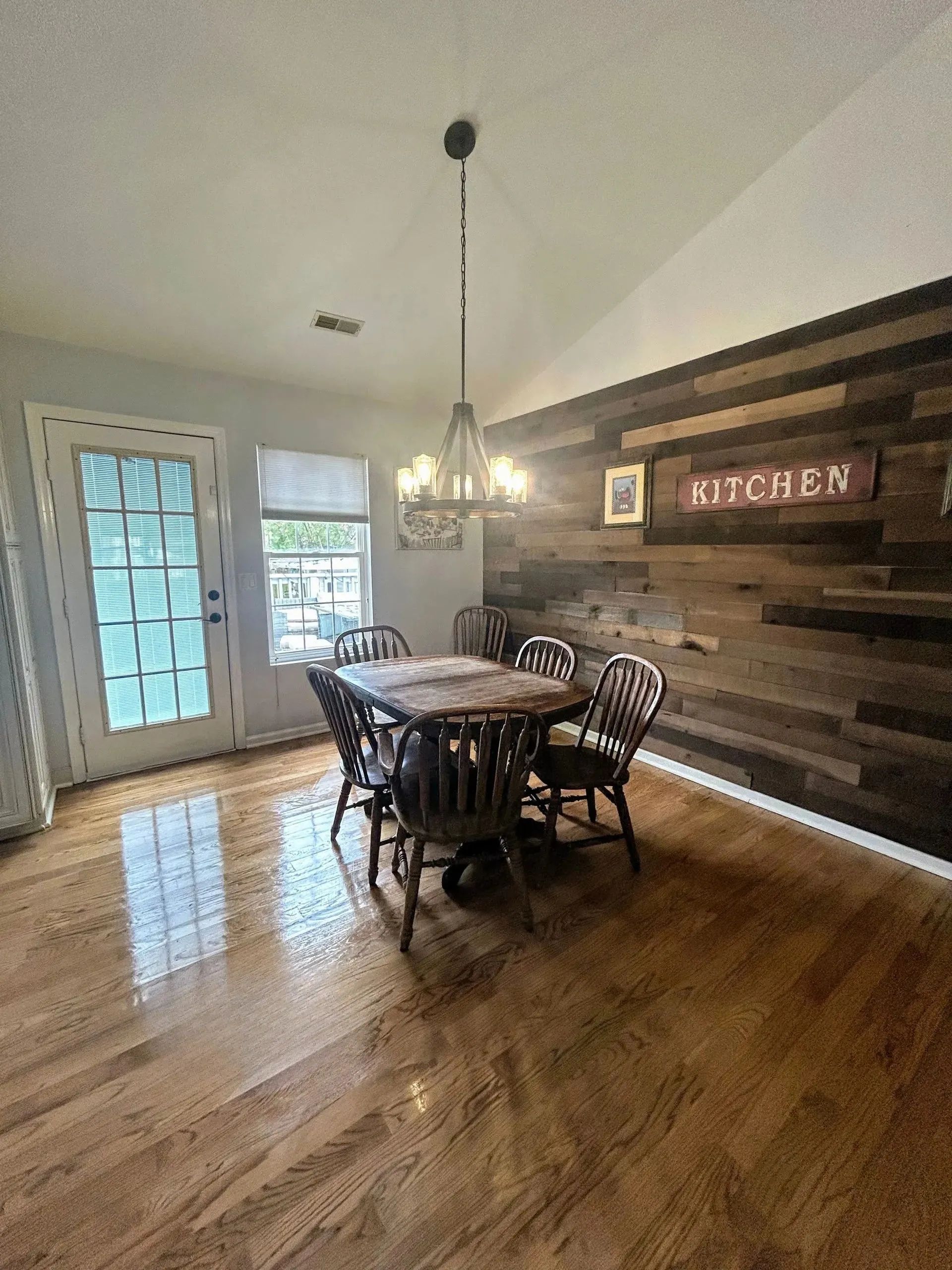 Dining room with wooden table and chairs, hardwood floor, and rustic plank accent wall with a “Kitchen” sign.