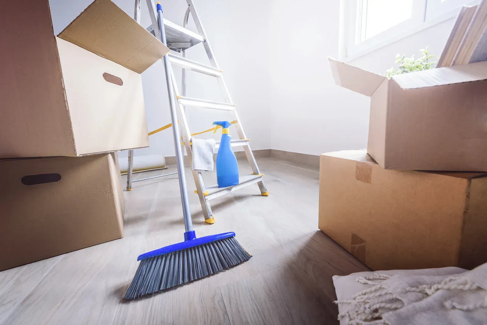 A blue broom stands in a room with cardboard moving boxes, a step ladder, and a cleaning spray bottle on a light floor.
