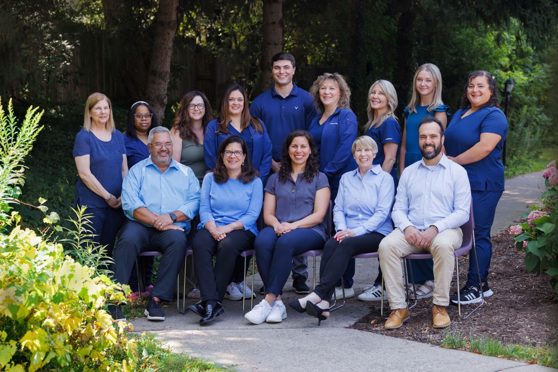 Group photo of a medical team in blue scrubs and shirts posing outside, smiling in front of foliage.