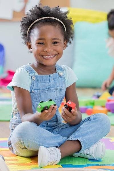 Girl in overalls sits cross-legged, holding toy cars, smiling brightly.