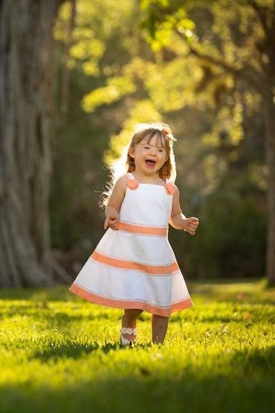 Smiling child in a white and orange dress running in a sunlit grassy park.