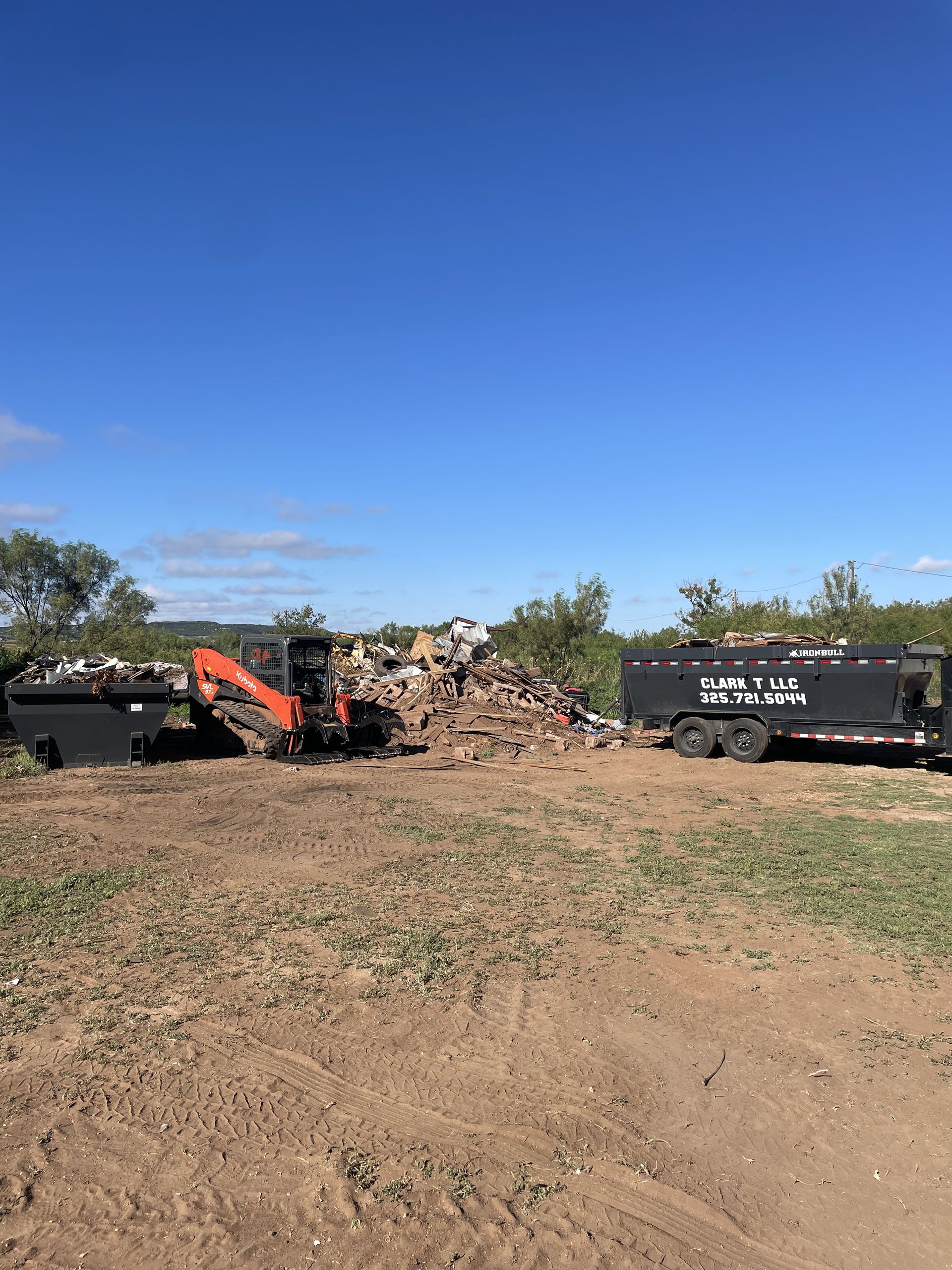 An excavator loads debris into dumpsters under a blue sky, on a dirt field.