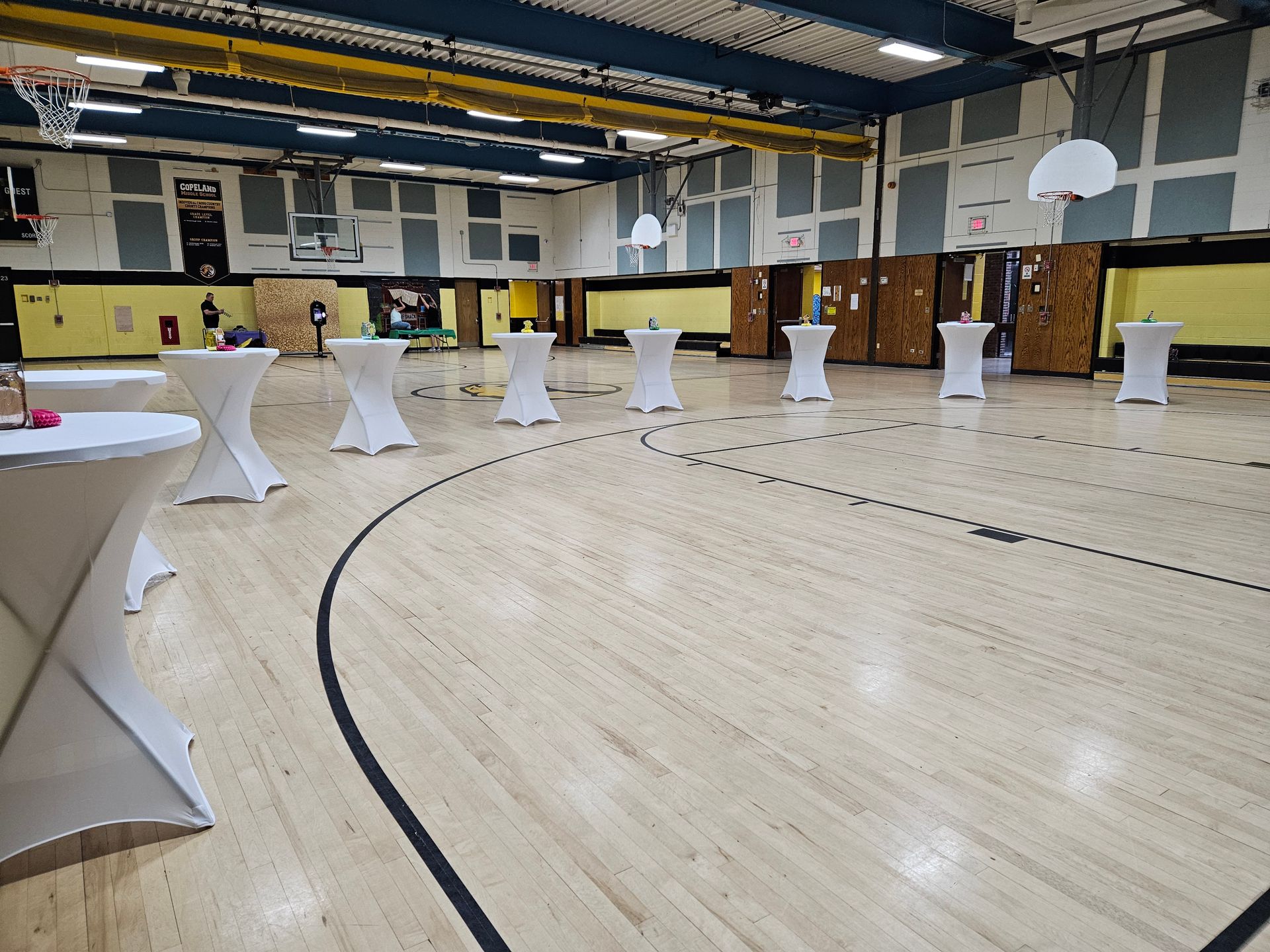 Basketball court set up for an event, with several white cocktail tables arranged. Yellow and blue walls, basketball hoops visible.