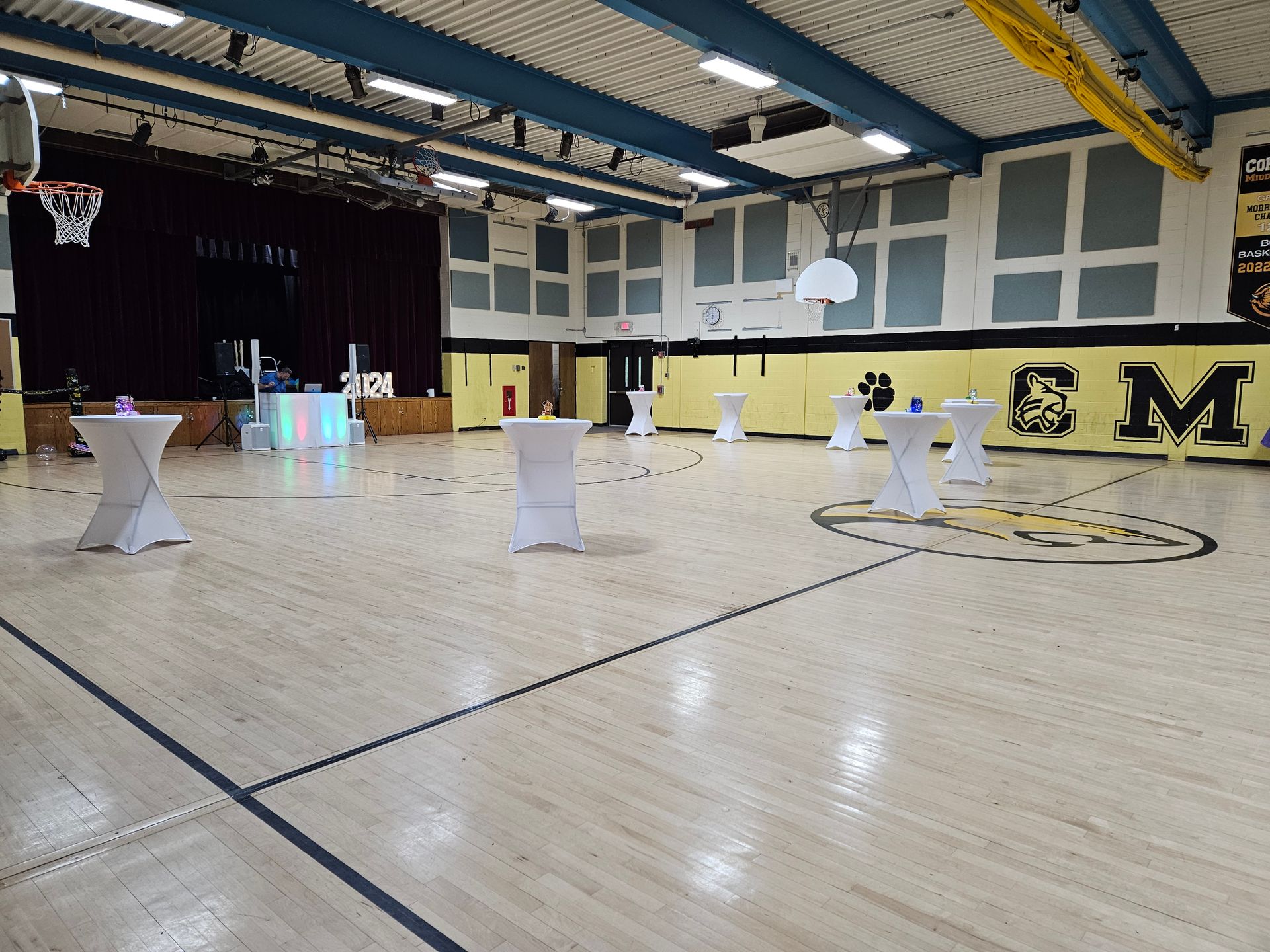 Gymnasium set up for an event, with white cocktail tables spread across the wooden floor. A stage and basketball hoops are visible.