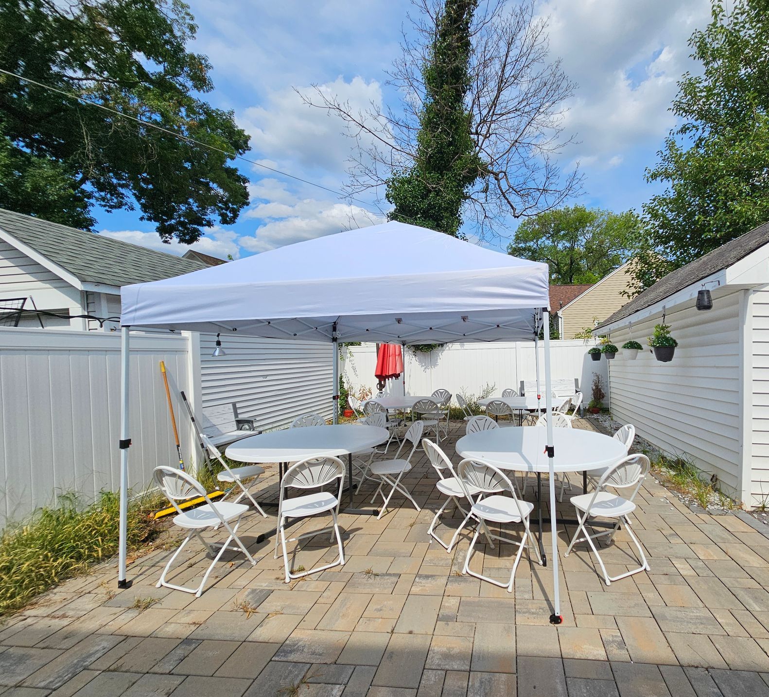 A white canopy shades round tables and white folding chairs on a patio. Trees and a white fence surround the space.