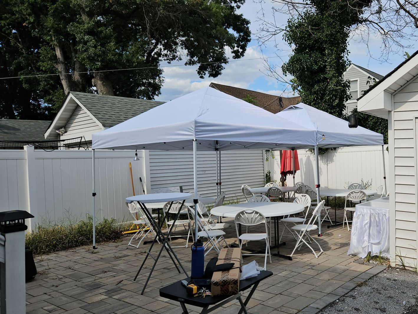 A backyard patio with a white canopy, tables, and chairs set up for an event. A white fence and buildings surround the area.