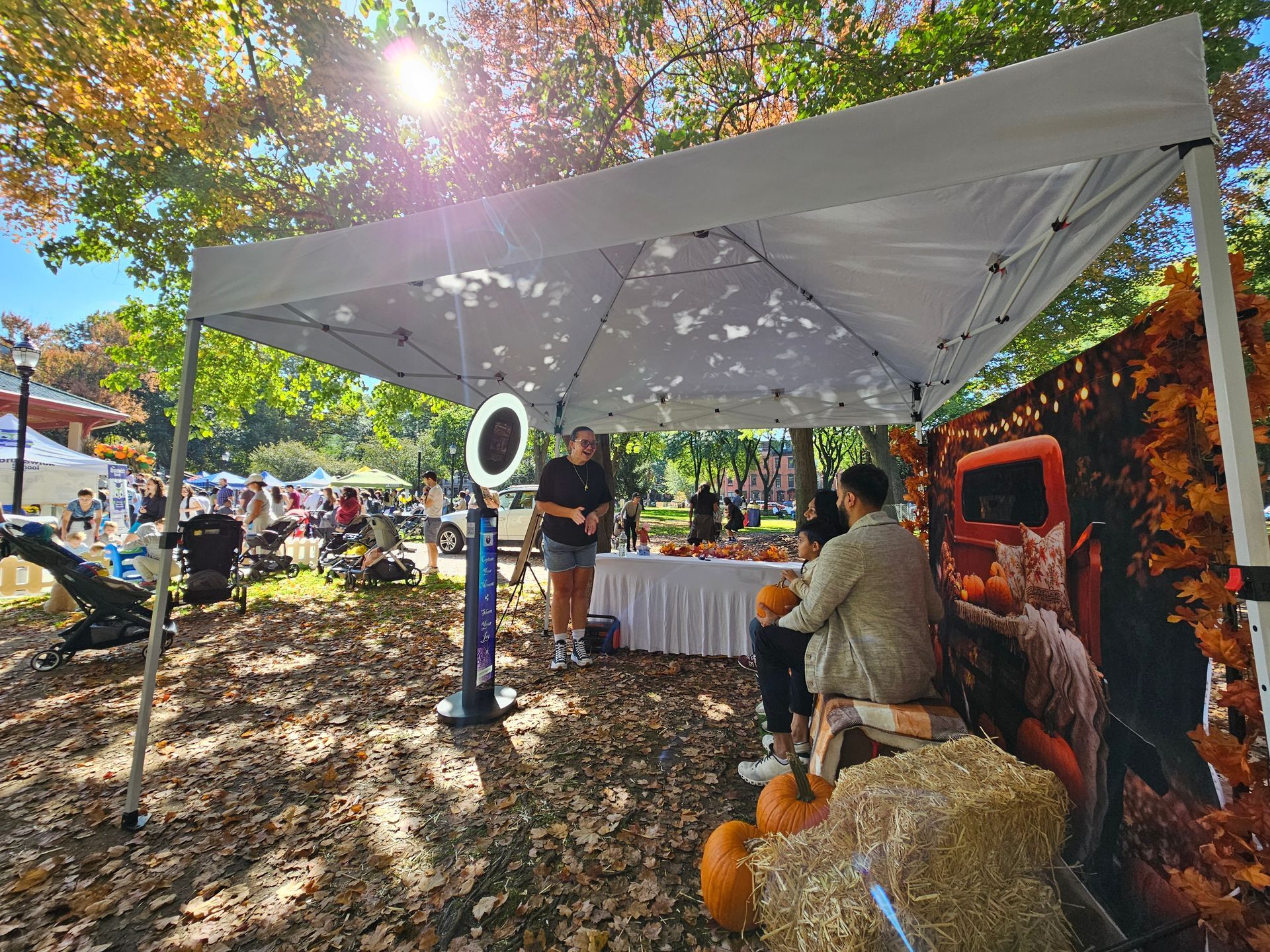 Photo booth at an outdoor autumn festival. People pose near a backdrop of a truck and hay bales, illuminated by ring light.