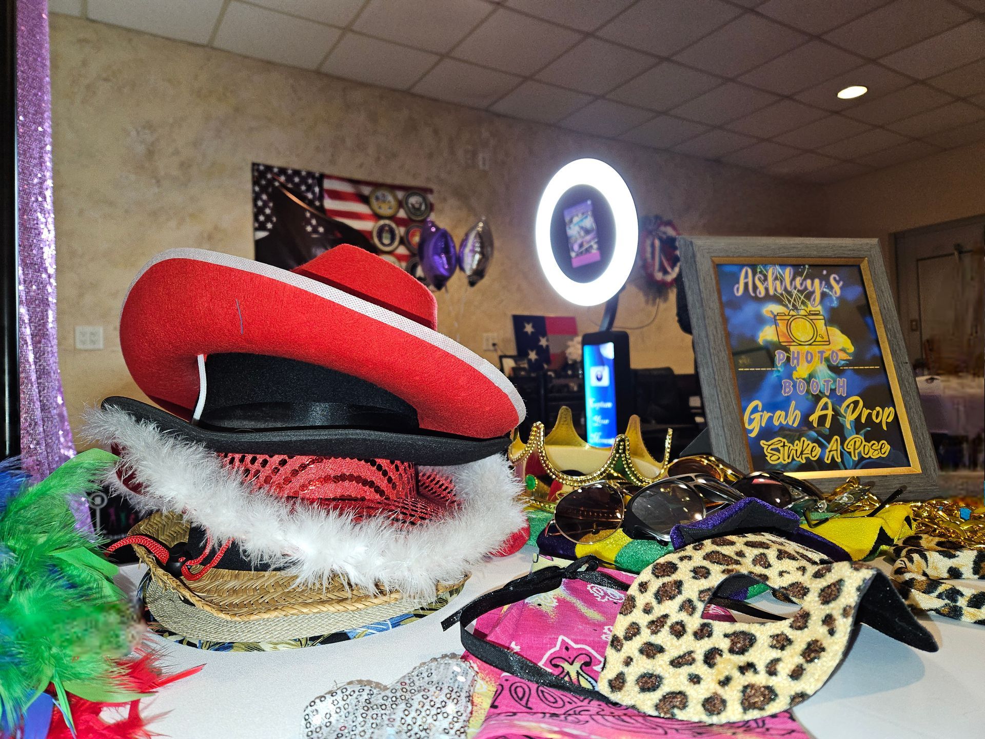 Photo booth props: hats, masks, and glasses on a table. A red hat is stacked on others. A lit ring light and sign are in the background.