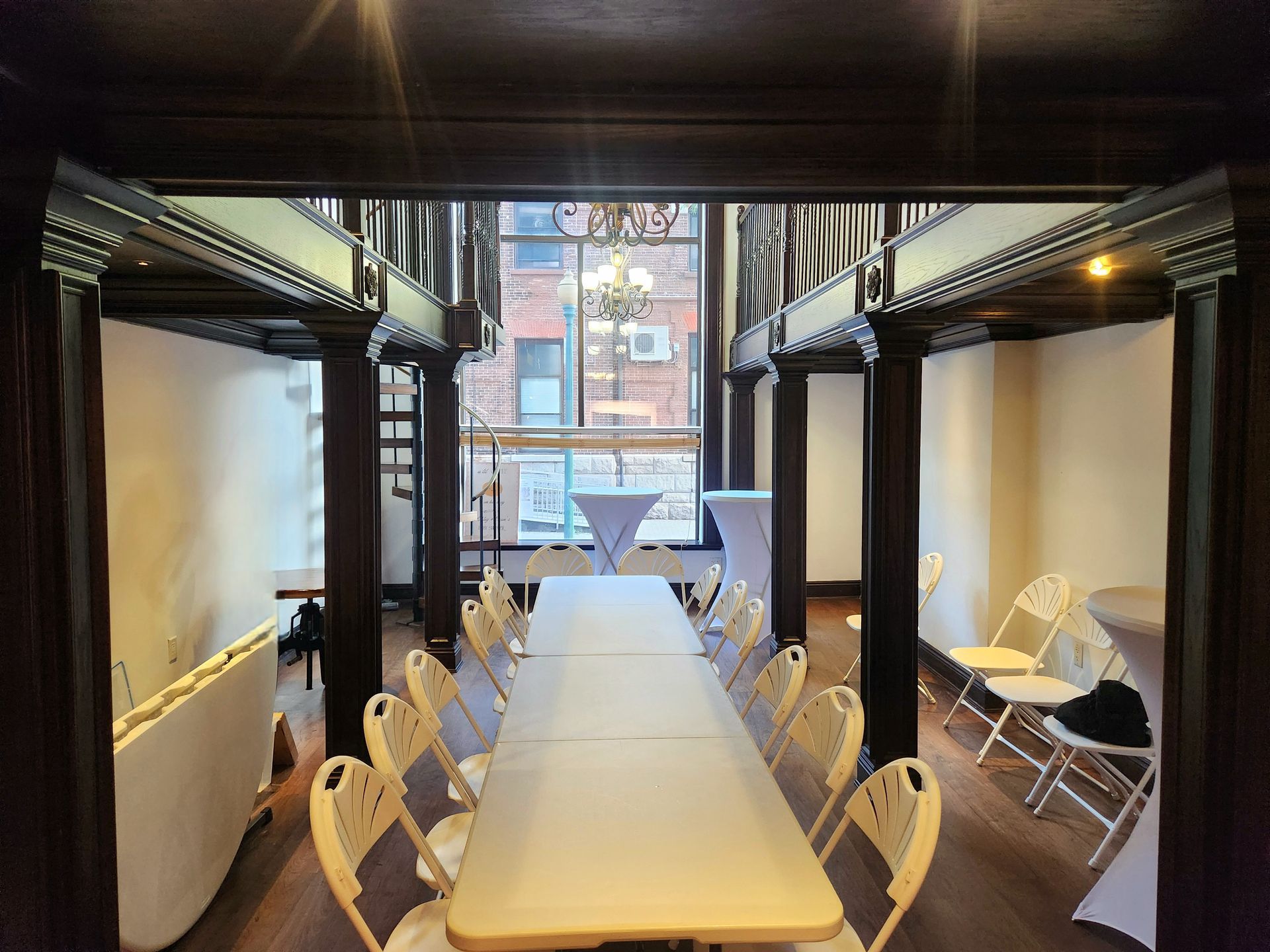 A long table set for a banquet in a room with dark wood beams, cream-colored chairs, and a window overlooking a city street.