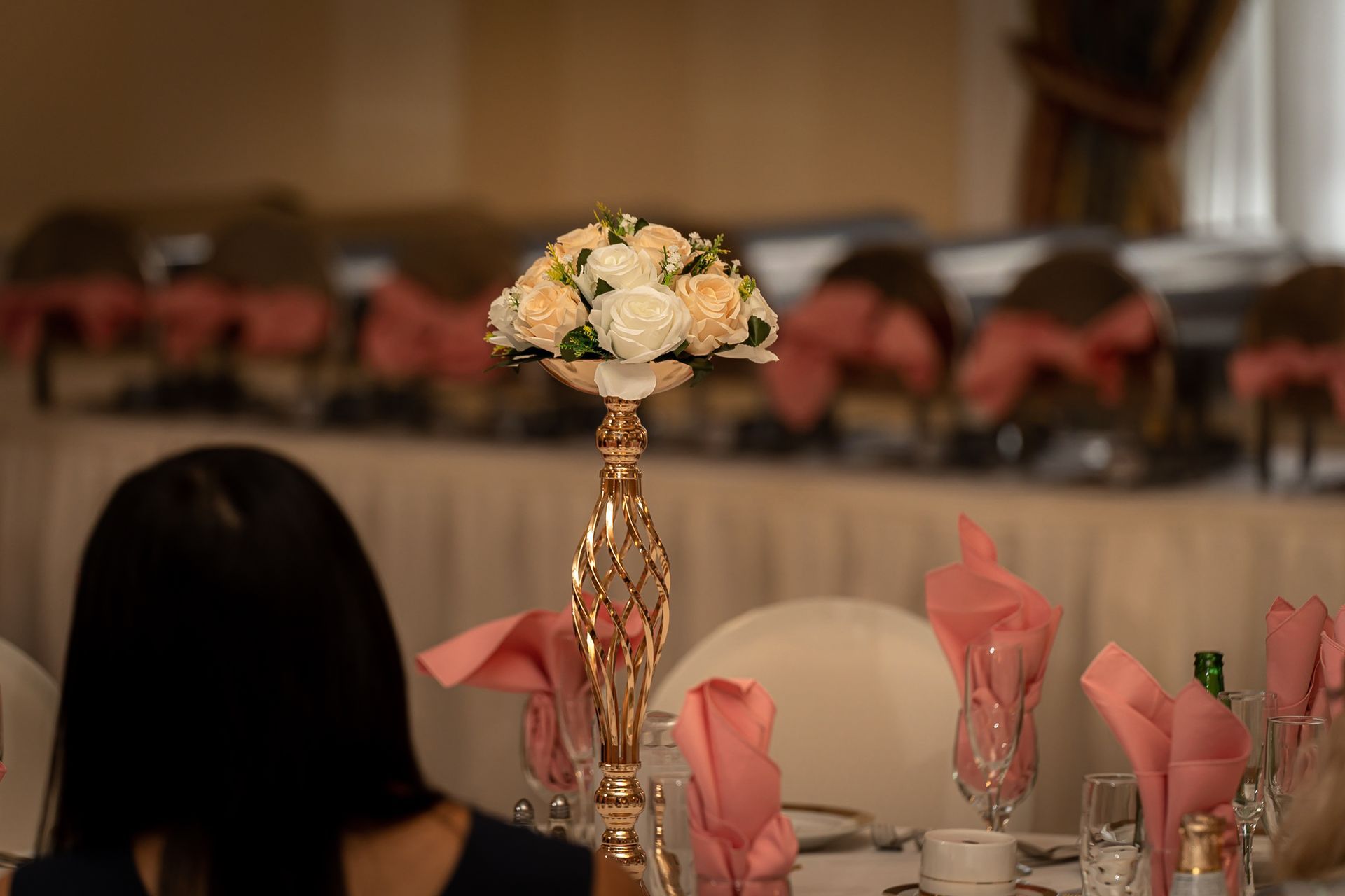 A centerpiece with a flower arrangement on a gold stand sits on a table set for a formal event. Pink napkins and other decorations are present.