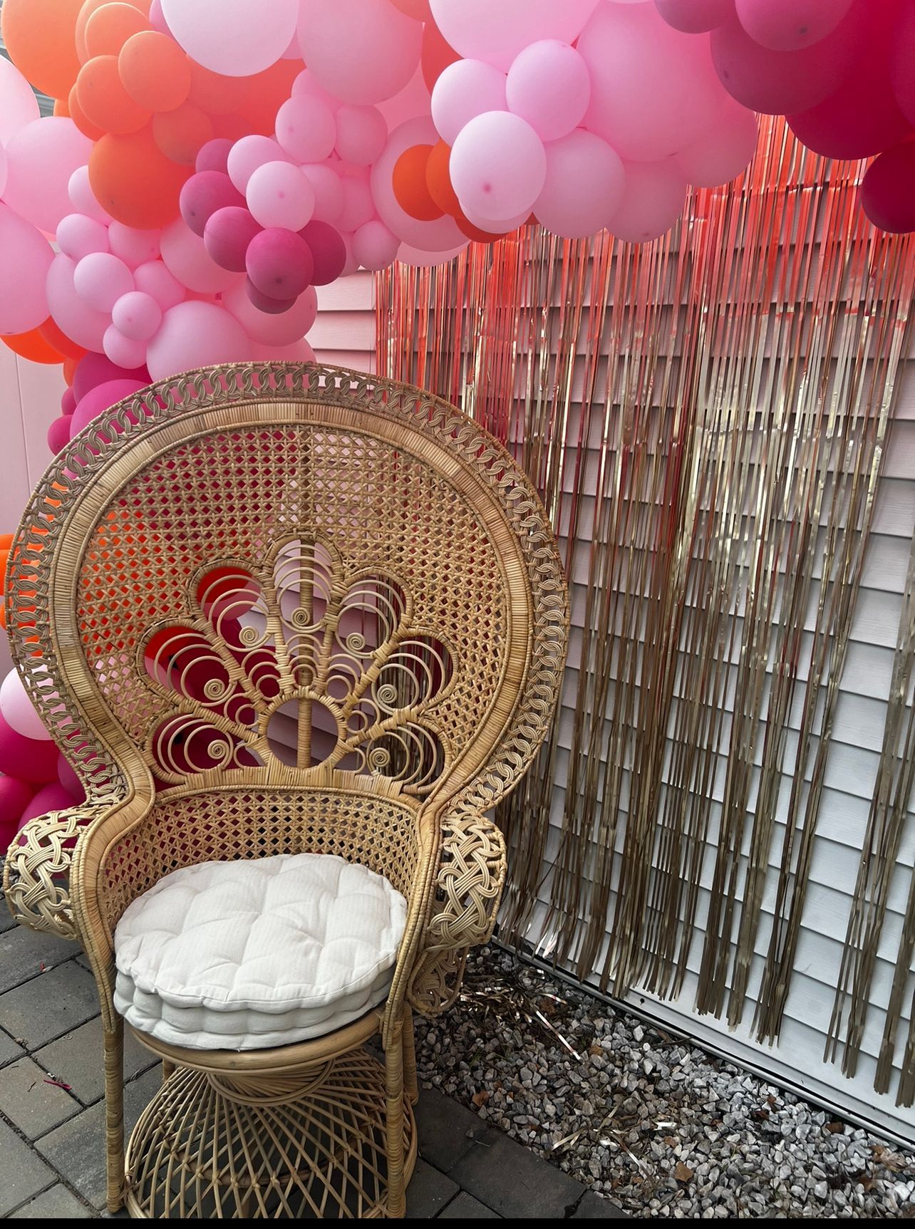 Wicker peacock chair with a white cushion beneath a cluster of pink and orange balloons, near a gold fringe backdrop.
