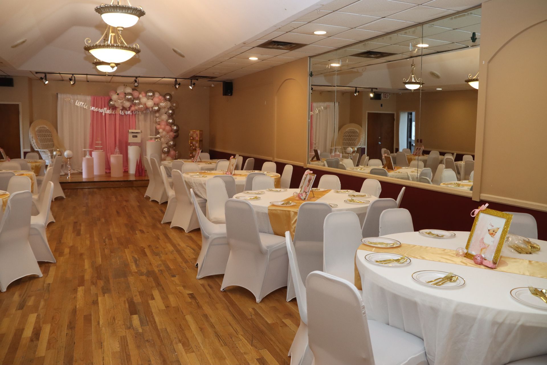An event space with round tables set for a celebration. White chair covers, gold runners, and pink decorations.  A stage and mirrored wall are visible.