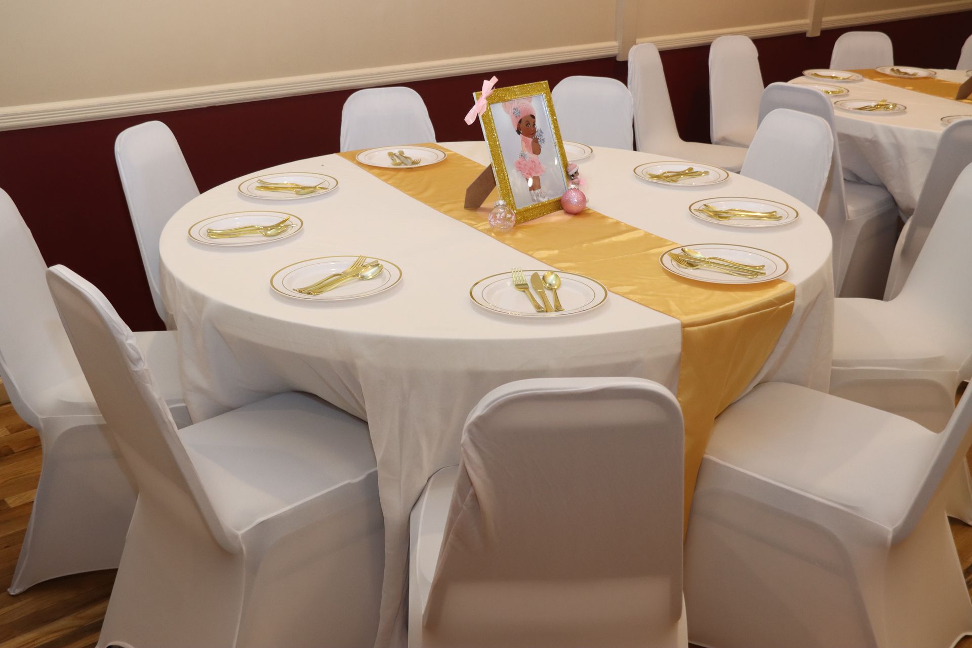 A round table set for a party, covered with a white cloth and gold runner, surrounded by white chair covers.