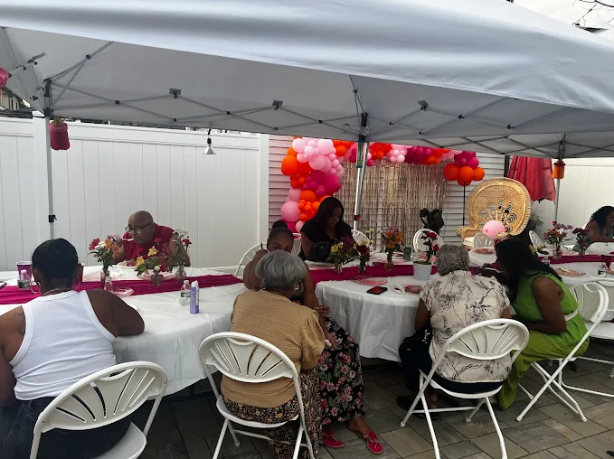 People seated at tables decorated for a party outdoors under a tent, with pink and white balloons.
