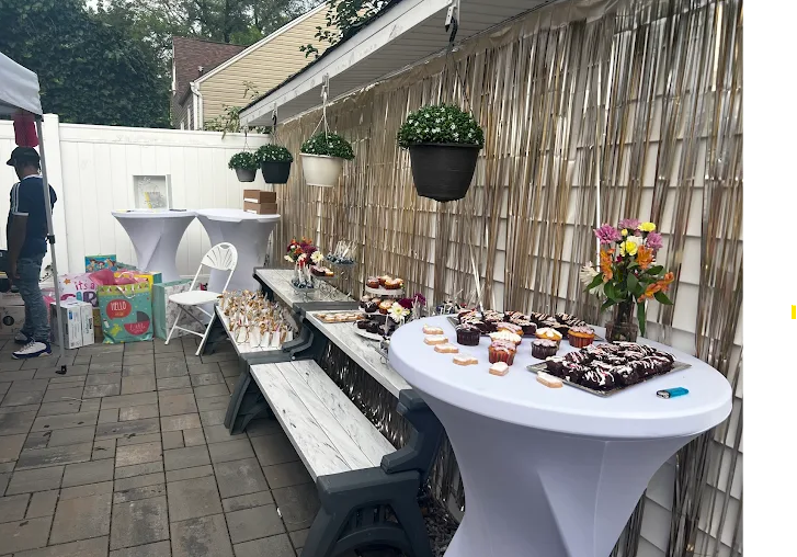 Backyard party scene with tables set for food and drinks, including a table covered in white fabric.  Plants hang from the wall.