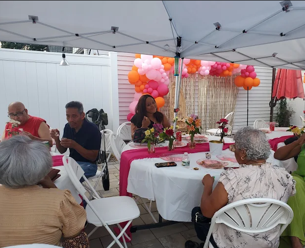 People seated at tables under a tent, likely at a party. Pink and orange balloons and decorations are in the background.