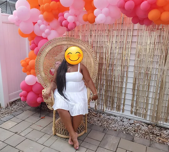 A person sits in a peacock chair, in front of a pink, orange, and gold balloon arch and gold fringe.