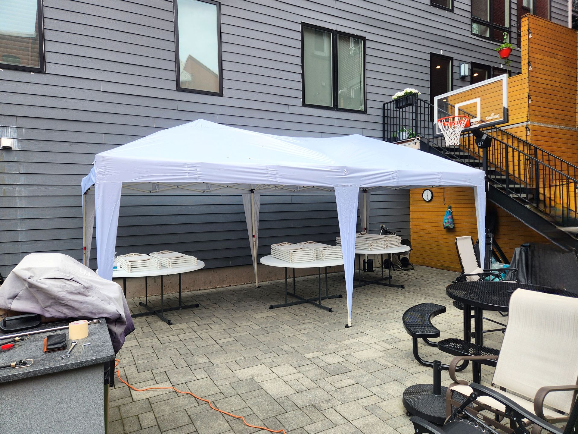 A white canopy covers round tables set up in an outdoor patio. A basketball hoop and stairs are visible in the background.