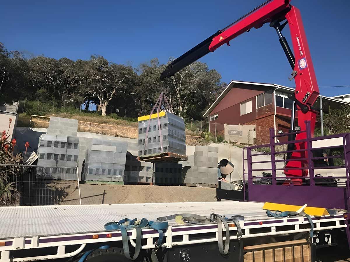 A Red Crane Is Lifting A Pallet Of Bricks From A Truck — Snap Crane Truck Hire in Port Macquarie, NSW