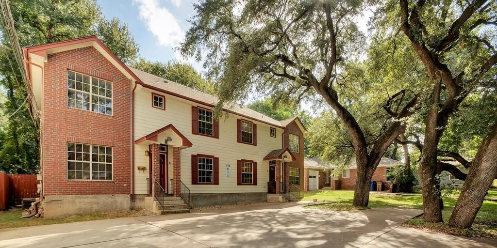 Two-story residential building with red brick and beige facade, shaded by large trees.