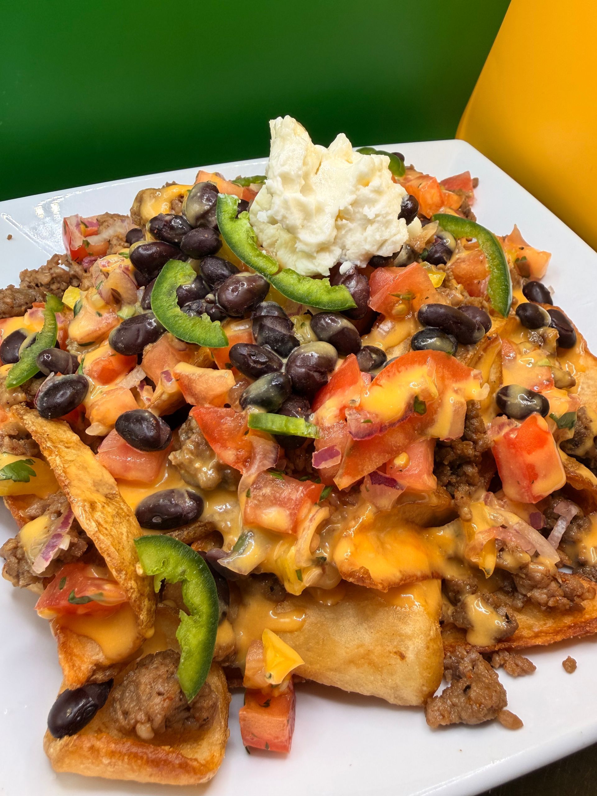 A plate of nachos topped with ground meat, black beans, diced tomatoes, sliced jalapeños, and a scoop of sour cream.