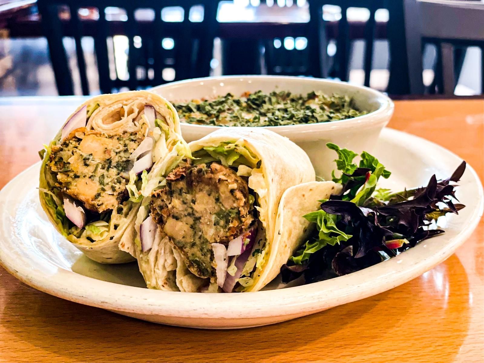 A falafel wrap cut in half on a white plate with a side of green dip and a small salad on a wooden table.
