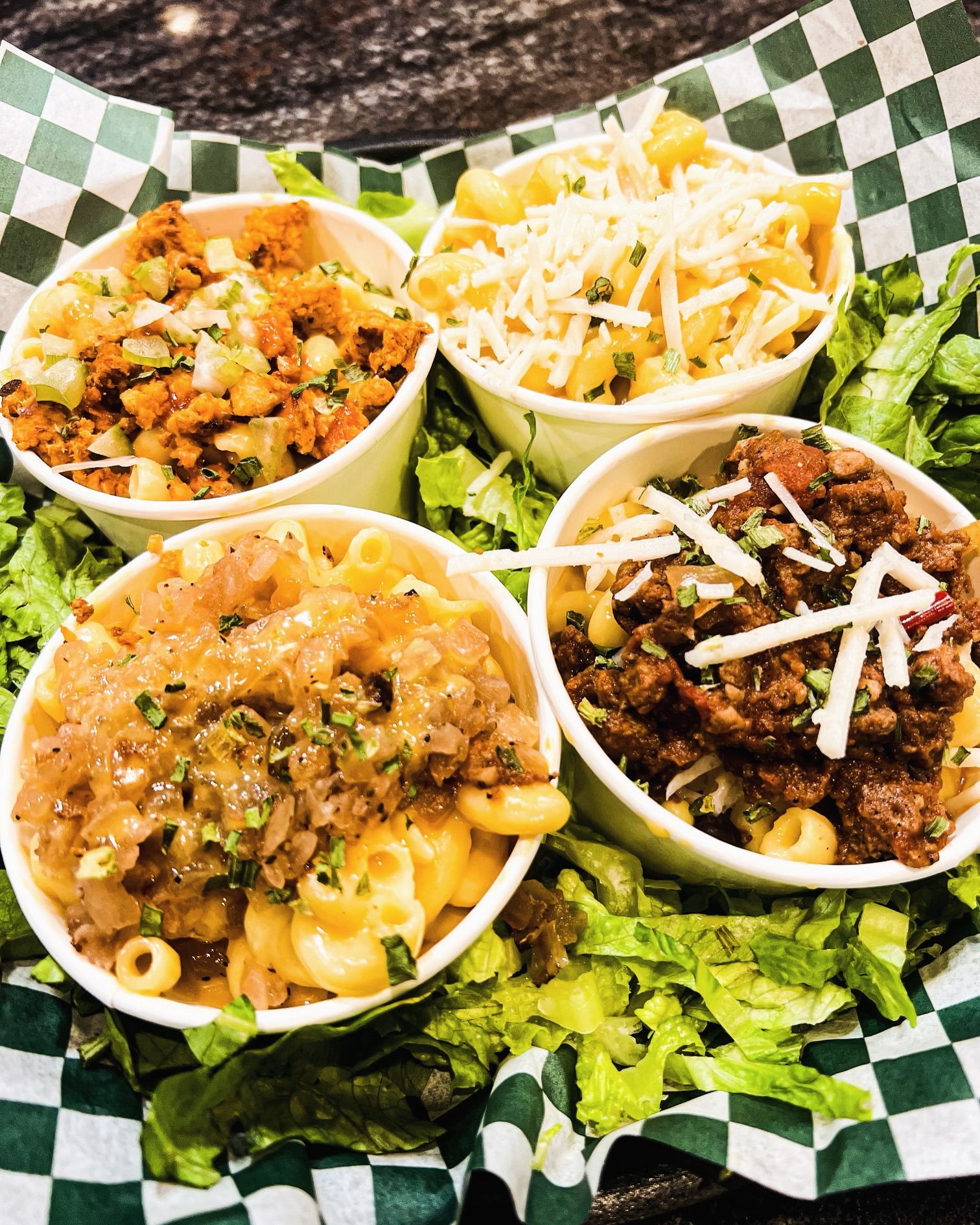 Four small white bowls of assorted macaroni and cheese topped with various ingredients, set in a green-checked basket.