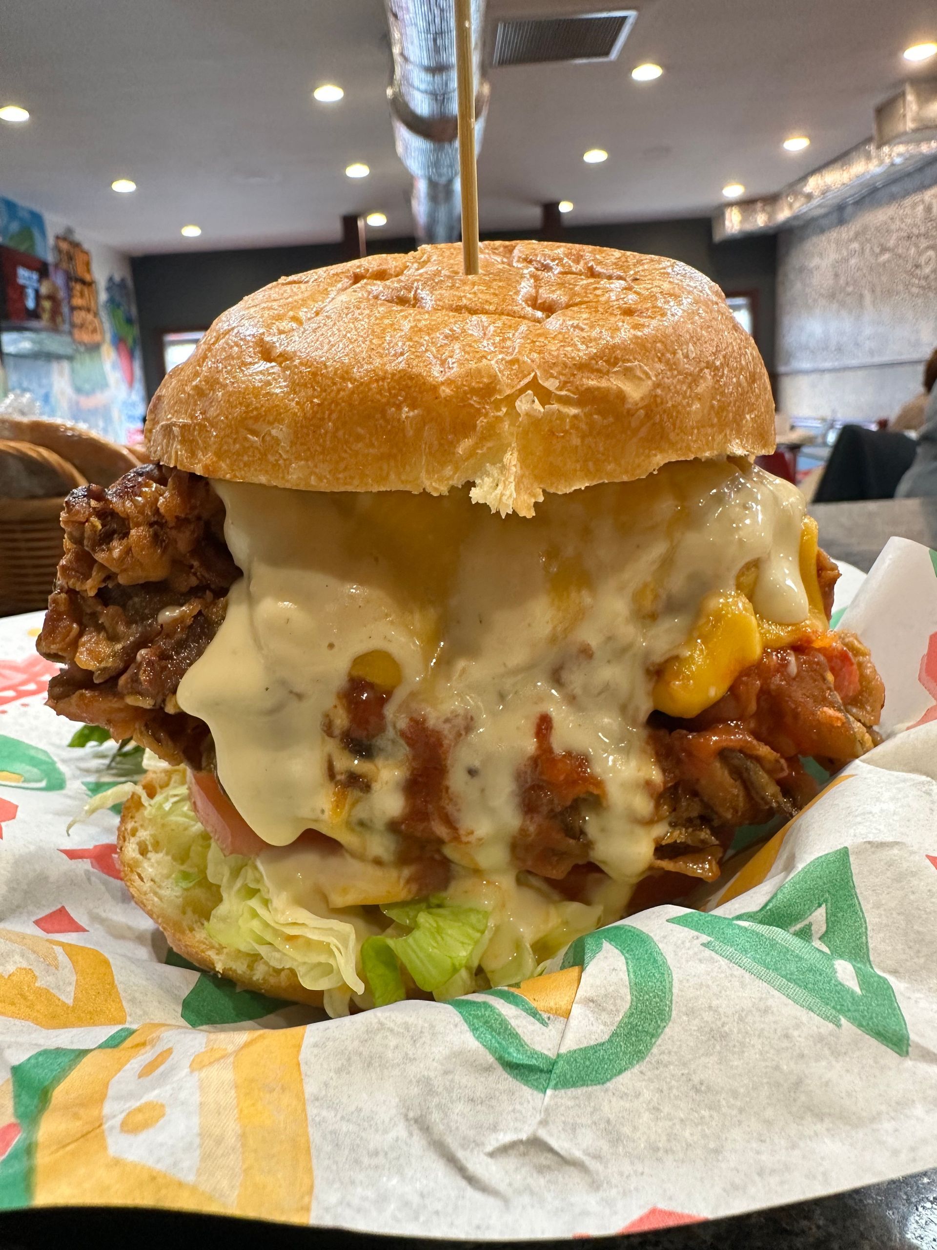 A large, double-patty burger with melted cheese and lettuce, served on patterned paper in a restaurant setting.