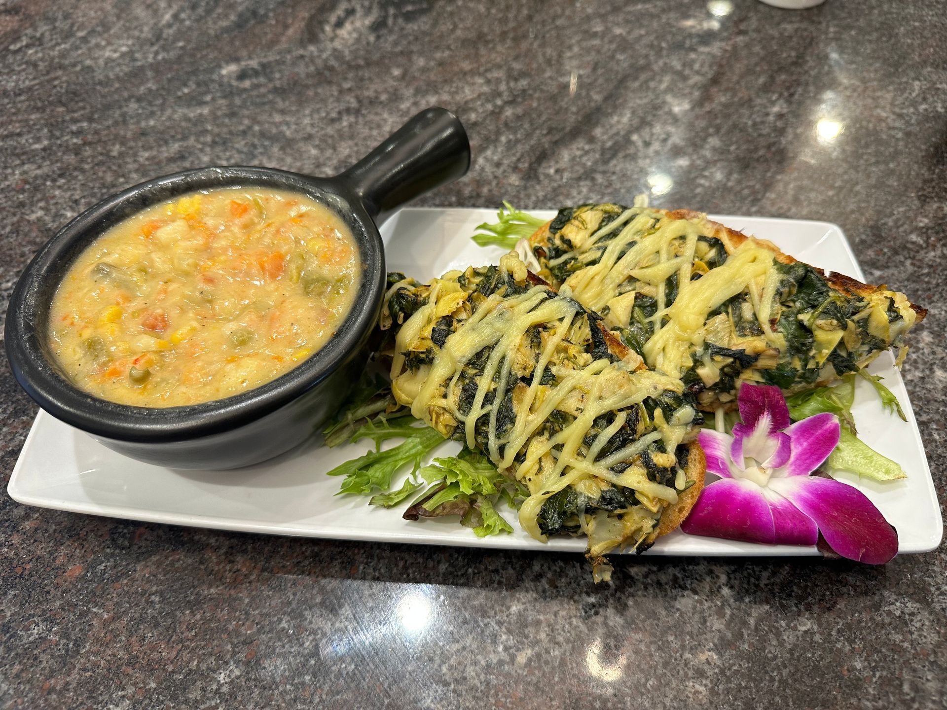 A small black bowl of vegetable soup served with two pieces of cheesy spinach toast on a plate with a purple orchid.