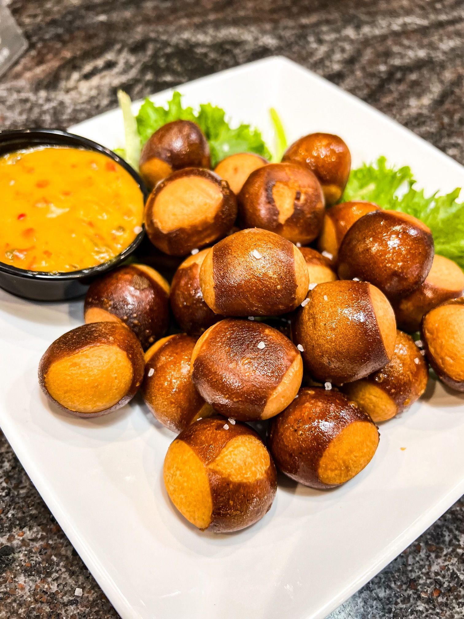 A plate of golden-brown pretzel bites sprinkled with salt, served with a side of dipping sauce on a bed of lettuce.