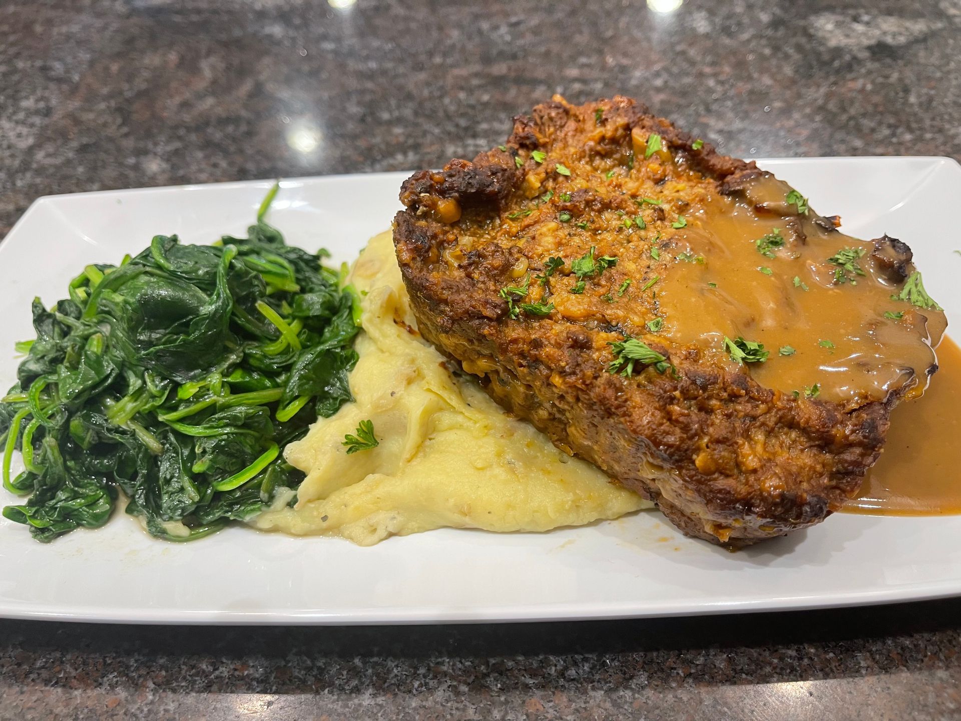 A white rectangular plate featuring a slice of meatloaf with brown gravy, a scoop of mashed potatoes, and sautéed spinach.