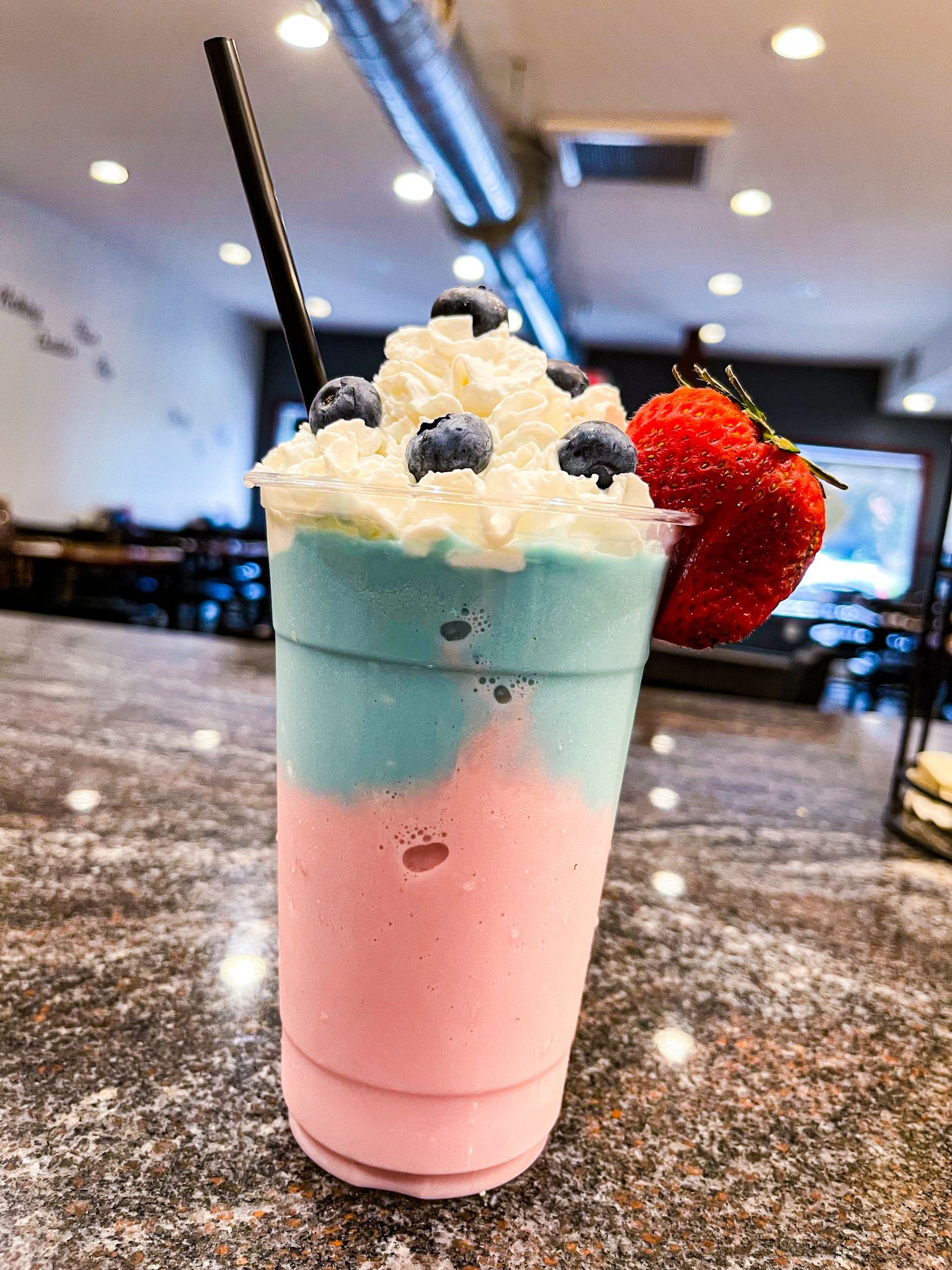A layered blue and pink milkshake topped with whipped cream, blueberries, and a fresh strawberry on a marble counter.