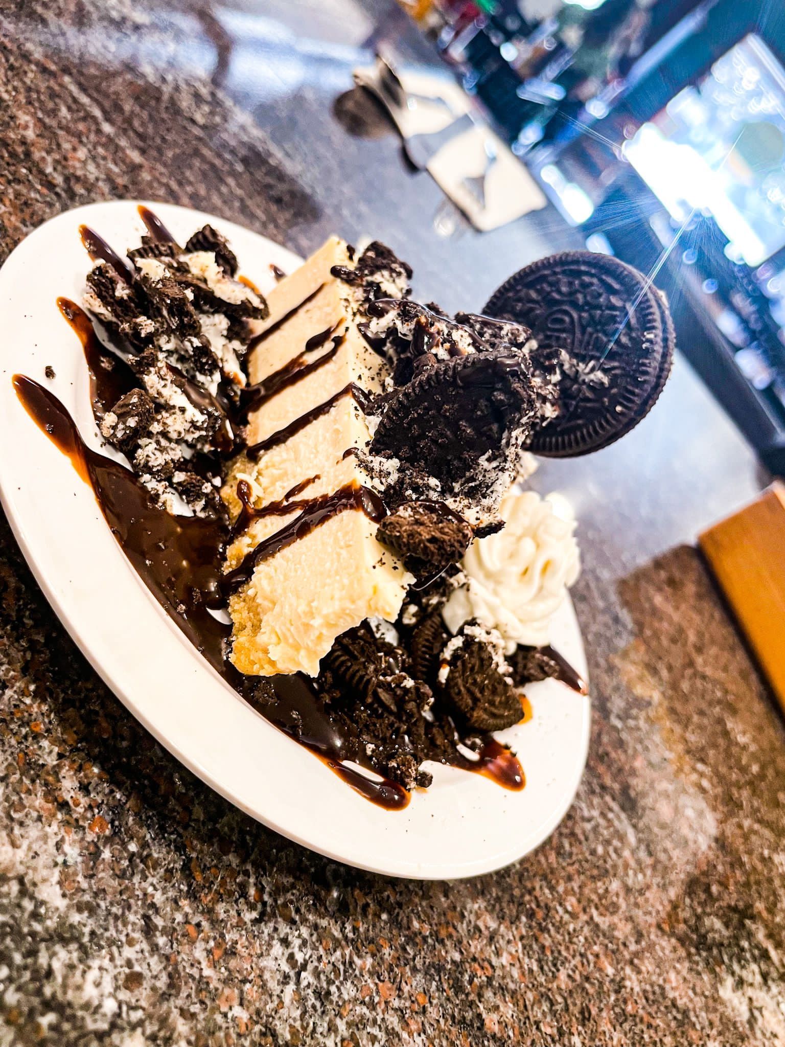 A slice of cheesecake topped with chocolate syrup, crushed cookies, whipped cream, and a whole Oreo on a white plate.