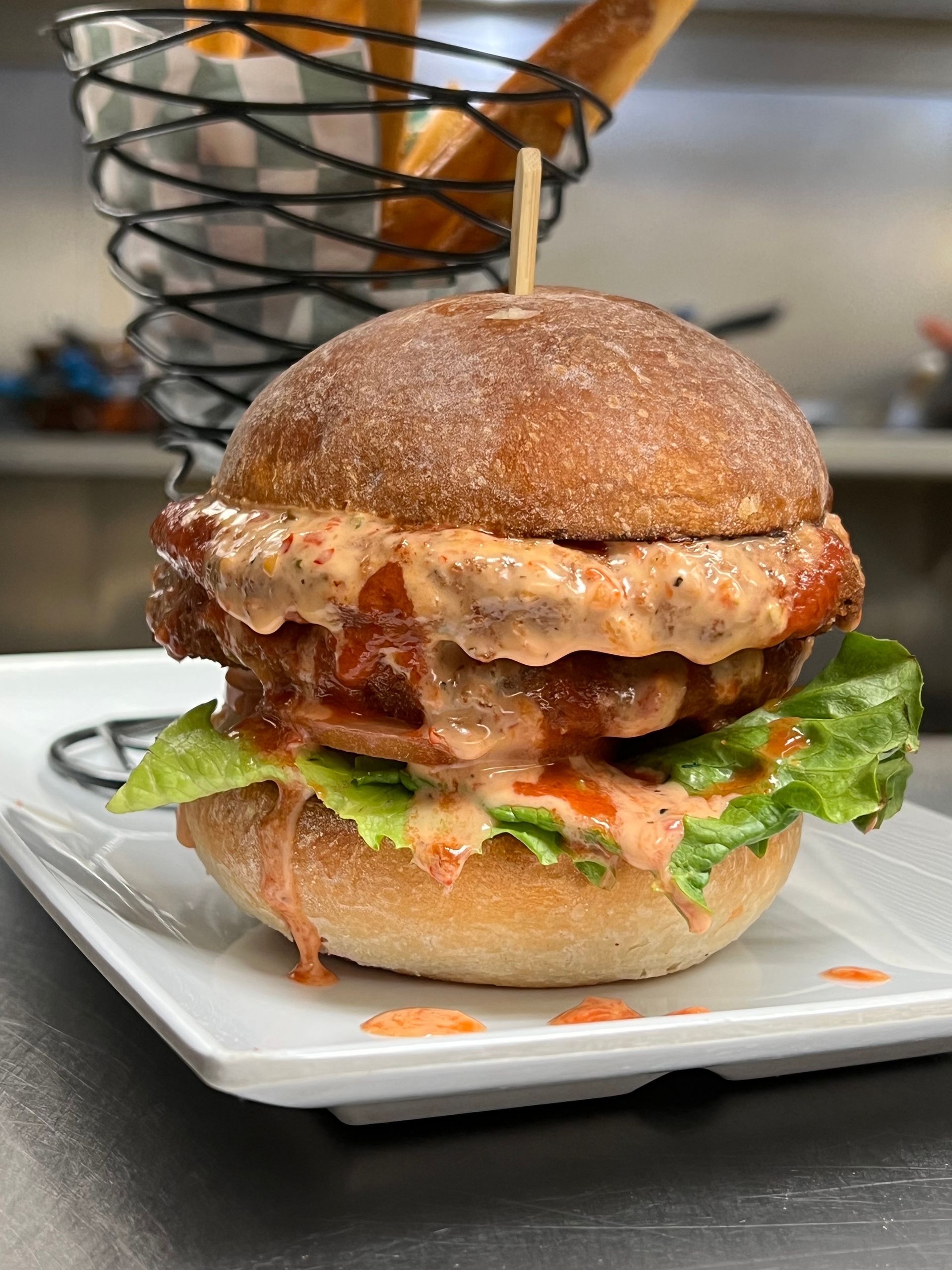 A chicken sandwich with lettuce and sauce on a bun, served on a white plate with a wire basket of fries in the background.