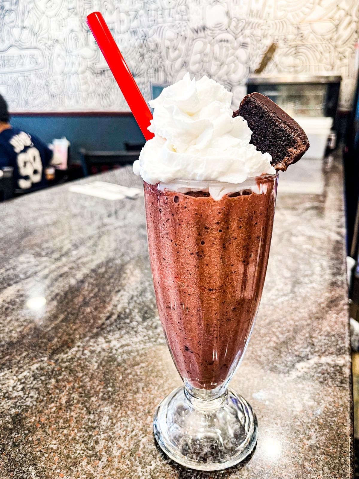 A chocolate milkshake topped with whipped cream and a brownie slice, served in a glass on a granite counter.