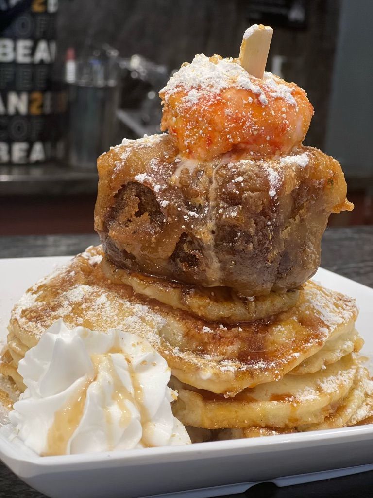 A stack of pancakes topped with a fried donut hole, orange garnish, powdered sugar, and whipped cream on a white plate.