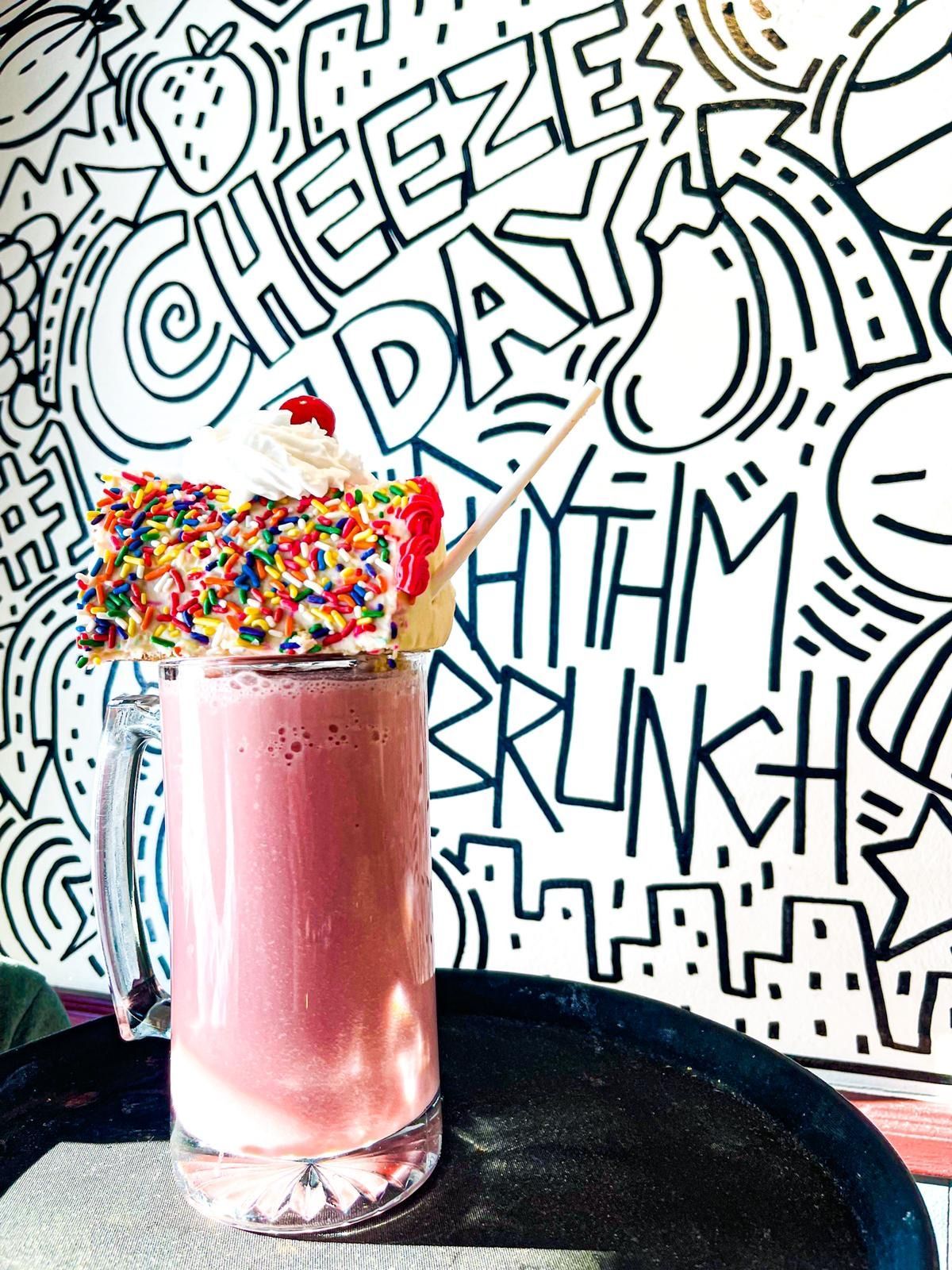 A pink milkshake topped with a large, sprinkle-covered cake slice, set against a black and white illustrated wall.