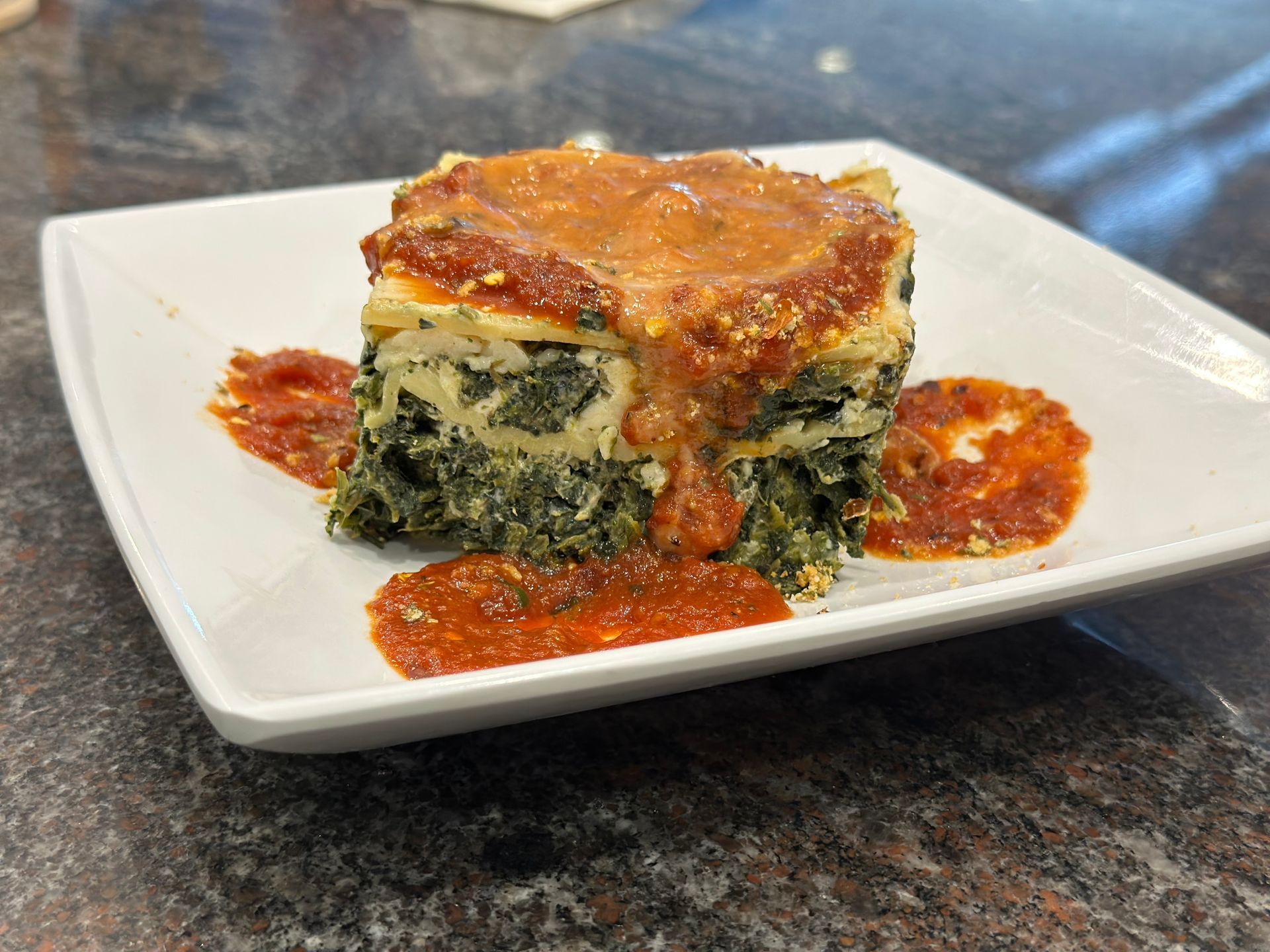 A square slice of spinach lasagna topped with red sauce, served on a white plate against a dark granite countertop.