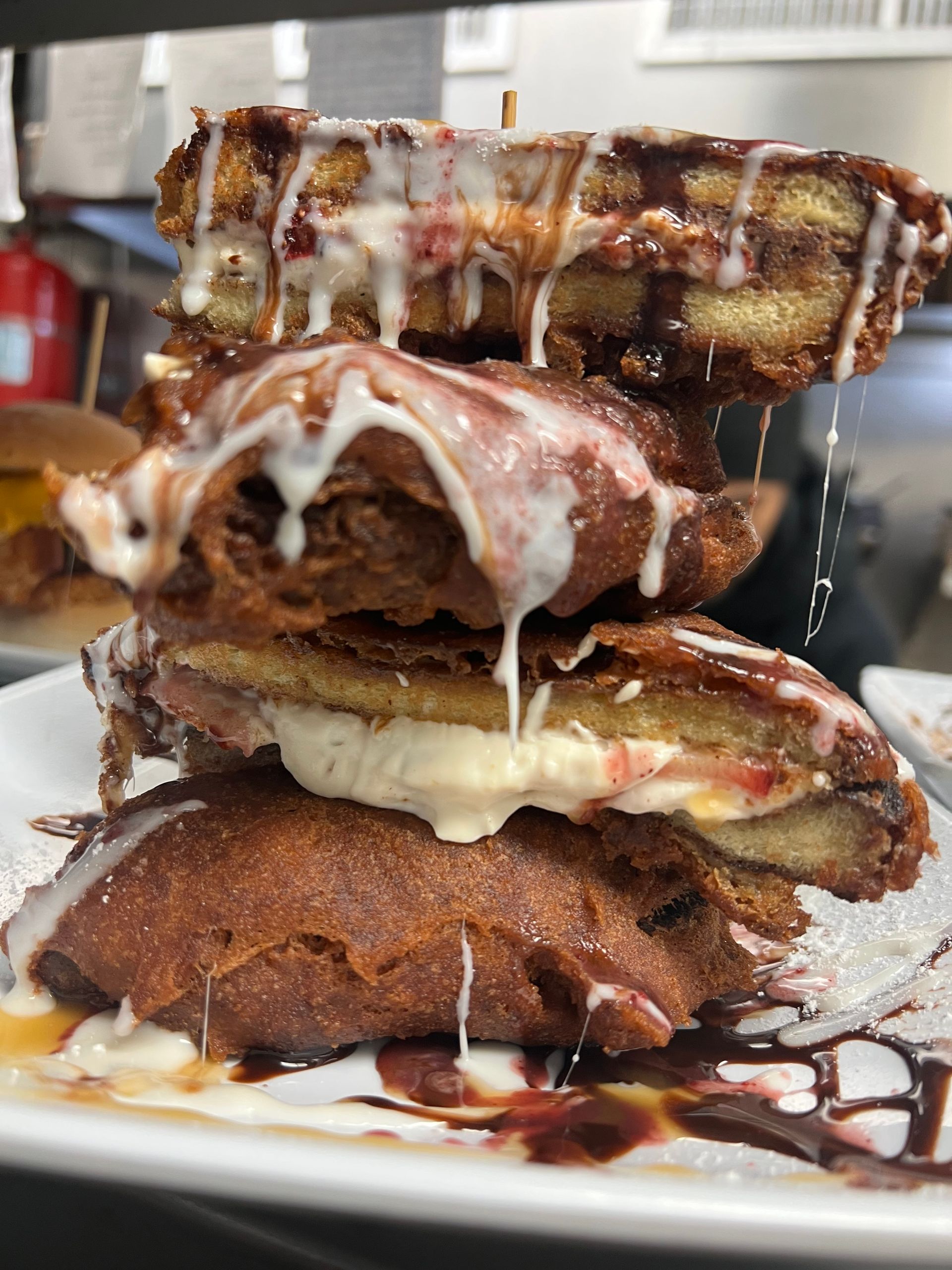 A tall stack of deep-fried, decadent dessert sandwiches drizzled with white icing and dark chocolate sauce on a plate.