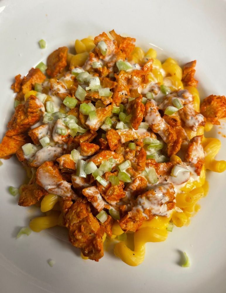 A bowl of macaroni and cheese topped with buffalo chicken, a drizzle of creamy sauce, and chopped celery.