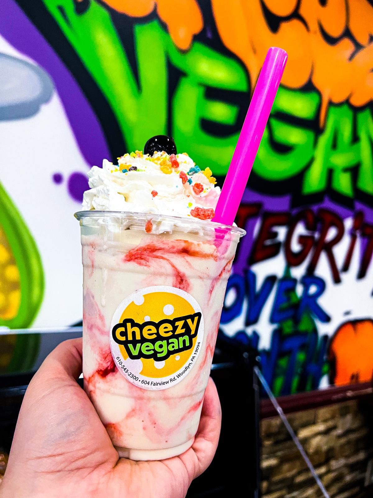 A hand holds a strawberry swirl vegan milkshake topped with whipped cream and sprinkles in front of graffiti art.